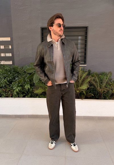 Hombre con gafas de sol, chaqueta de cuero sobre un jersey beige, pantalones oscuros y zapatillas blancas, de pie sobre un suelo de baldosas cerca de plantas.