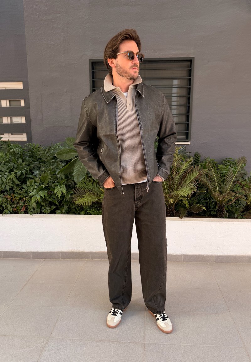 Homme portant des lunettes de soleil, une veste en cuir sur un pull beige, un pantalon foncé et des baskets blanches, debout sur un sol carrelé près des plantes.