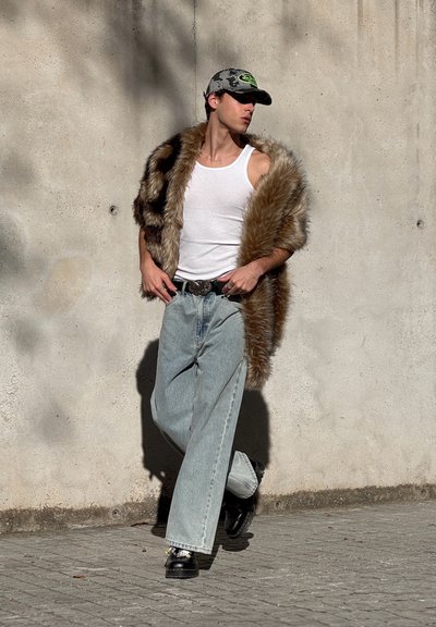 Abrigo de piel sintética con patrones marrón y negro, camiseta sin mangas blanca, jeans ligeros de color azul claro, zapatos negros con detalles plateados y una gorra de béisbol camuflada.