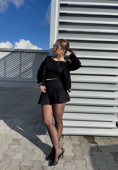 Chaqueta bomber negra con rayas blancas, crop top negro, falda plizada negra, medias negras transparentes y tacones altos negros sobre un fondo gris.