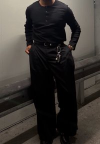 Chemise noire à manches longues côtelée avec patte de boutonnage, associée à un pantalon noir à jambes larges. Une ceinture noire avec des clés attachées à la taille.