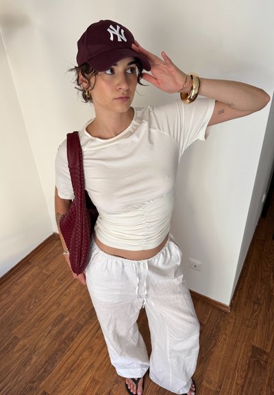 Top blanco corto con lados fruncidos, combinado con pantalones blancos sueltos. Bolso de mano rojo oscuro con diseño trenzado y una gorra burdeos con logo blanco.