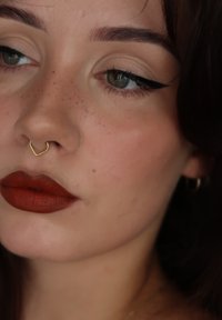 Gul hjärtformad septumsmak, djup röd matt läppstift, naturlig hudton med fräknar, väldefinierade ögonbryn och vingad eyeliner.