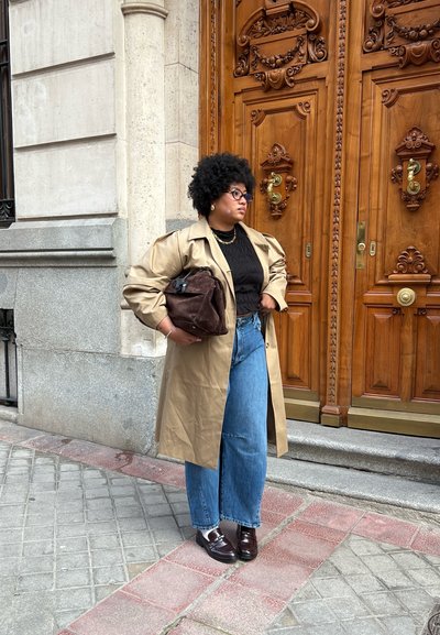 Un abrigo trench beige sobre una blusa negra texturizada, jeans anchos azules y zapatos marrones. Lleva un bolso marrón oscuro y está de pie cerca de puertas de madera.