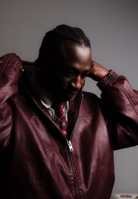 Homme aux cheveux tressés portant une veste en cuir rouge, ajustant le col, avec une cravate rouge à motifs et des colliers en couches sur fond gris.