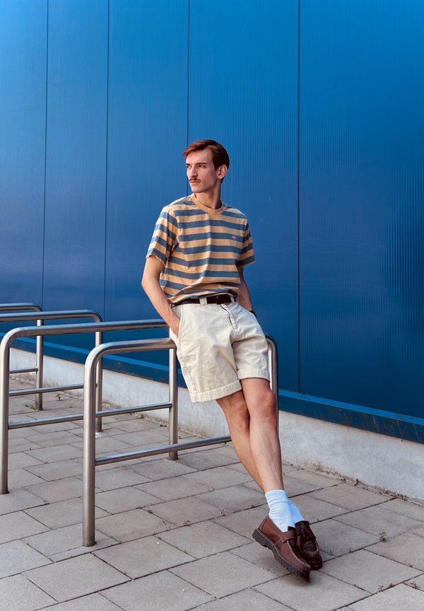Jeune homme en chemise rayée et short beige appuyé contre une rampe métallique avec un mur bleu en arrière-plan, mains dans les poches, regardant sur le côté.