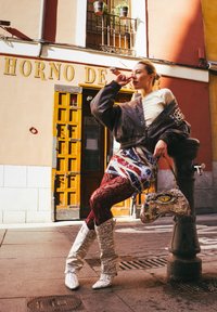 Braune Bomberjacke, gemustertes Kleid mit Union Jack, rote Spitzenstrumpfhose, silberne kniehohe Stiefel und eine Handtasche mit Reptilienmuster. Städtische Umgebung.