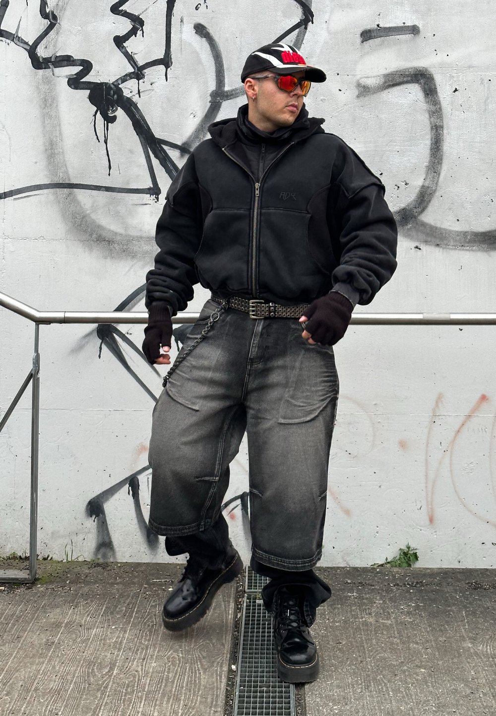 Giacca nera con zip, pantaloni cargo grigi e larghi, guanti senza dita e stivali platform neri. Gli accessori includono un cappello e occhiali da sole.