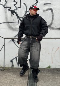 Giacca nera con zip, pantaloni cargo grigi e larghi, guanti senza dita e stivali platform neri. Gli accessori includono un cappello e occhiali da sole.