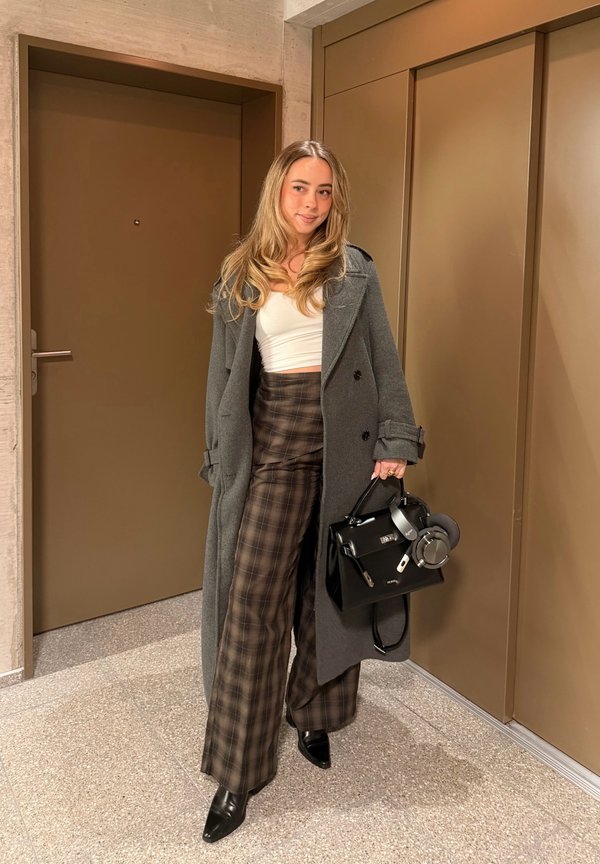 Ženska, ki stoji v prostoru, oblečena v dolg sivi plašč, bel kratek top, široke karo hlače, črne škornje in drži črno torbico s slušalkami.