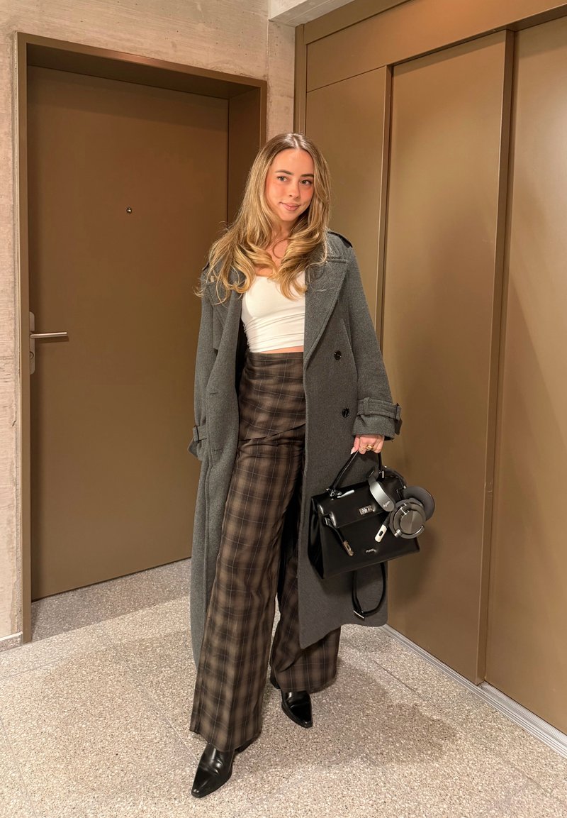 Γυναίκα που στέκεται σε εσωτερικό χώρο φορώντας μακρύ γκρι παλτό, λευκό κοντό τοπ, καρό παντελόνι με φαρδιά μπατζάκια, μαύρες μπότες και κρατώντας μια μαύρη τσάντα χειρός με ακουστικά.