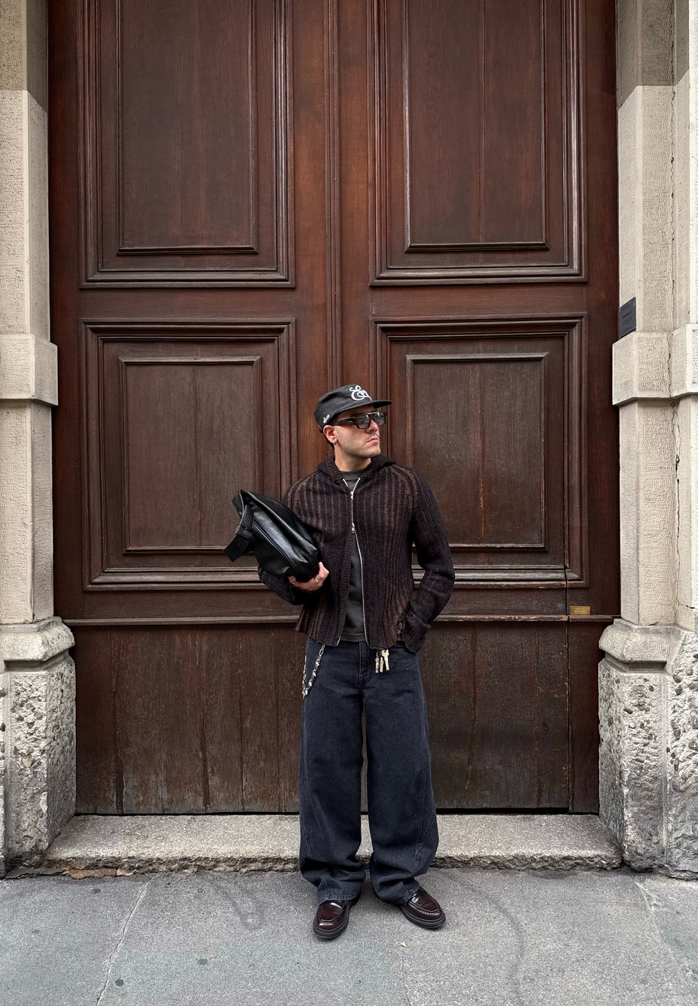 Giacca nera a coste con zip sopra una camicia scura, jeans larghi scuri e un cappellino nero. Tiene una borsa nera davanti a uno sfondo di porta in legno.
