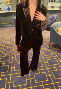 Osoba oblékající tmavě hnědý sametový oblek se širokými kalhotami, držící malou kabelku, stojící na modrém koberci se žlutými geometrickými vzory.