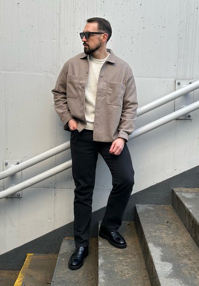 Chaqueta de camisa de lana marrón claro con dos bolsillos frontales, llevada sobre un suéter de punto crema y pantalones negros, con zapatos de cuero negros.