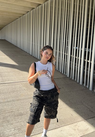 Top blanco sin mangas, pantalones cortos tipo cargo de camuflaje, bolsa negra de hombro, sosteniendo unas gafas, de pie en un pasillo con barandillas blancas verticales.