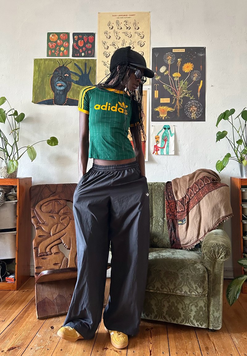 Personne portant un t-shirt vert Adidas, un pantalon noir, une casquette et des lunettes de soleil se tient dans un salon avec des plantes, des œuvres d'art et un fauteuil à motifs.