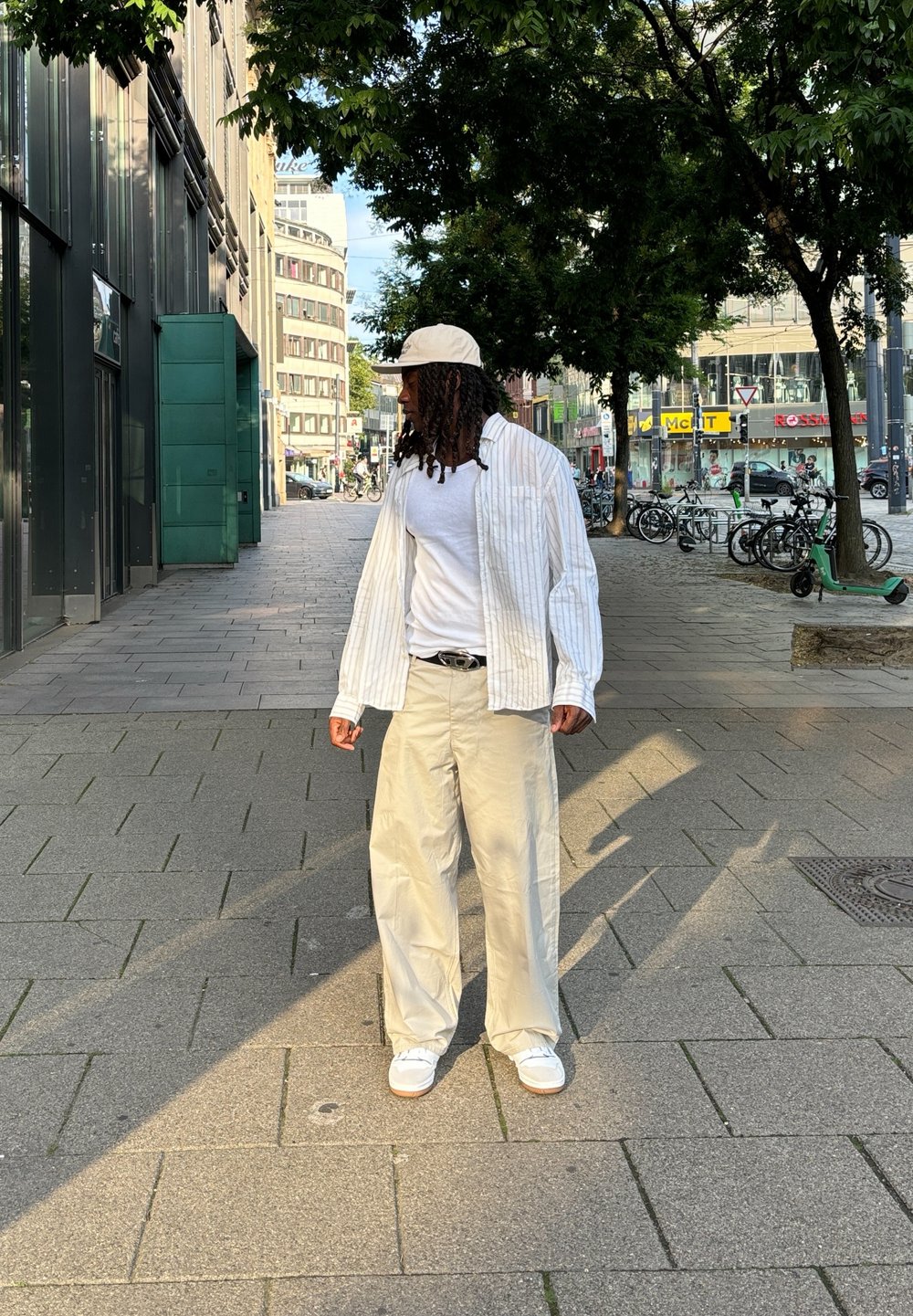 Camicia leggera a righe bianche sopra una maglietta bianca, pantaloni beige a gamba larga e sneakers bianche, su uno sfondo urbano.