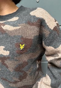 Sweter w kamuflażowym wzorze w odcieniach szarości, brązu i beżu. Na piersi znajduje się żółte haftowane logo orła. Miękka, dzianinowa faktura.