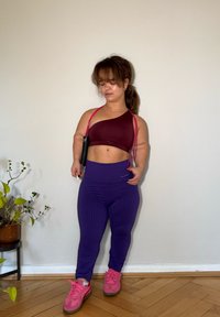 Sostegno sportivo bordeaux a spalla unica, leggings viola a trama, e scarpe da ginnastica rosa, su sfondo neutro con una pianta in un vaso accanto.