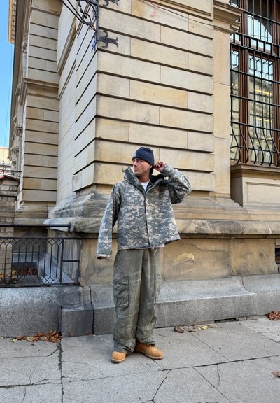 Chaqueta exterior de camuflaje con tonos grises y verdes, pantalones cargo grises, botas marrones; de pie junto a una pared de piedra texturizada.