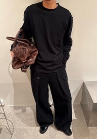 Chemise noire à manches longues avec une poche poitrine, associée à un pantalon noir ample avec de gros boutons. Un sac en cuir marron est tenu en main.