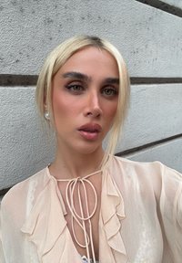 Ljus persikofärgad blus med volang framtill och knytning, genomskinligt tyg, kompletterad med pärlörhängen och rakt blont hår mot en neutral bakgrund.