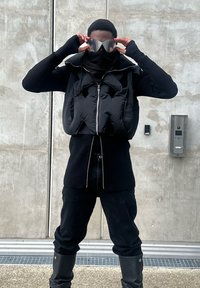 Black padded vest with a high collar worn over a black ribbed top. Paired with black trousers and knee-high boots. Holding reflective sunglasses; textured background.