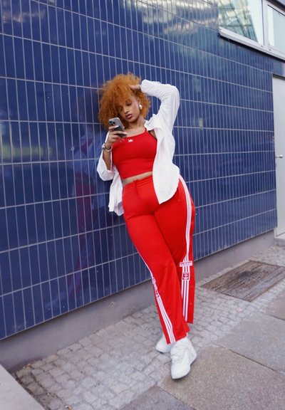 Top atlético rojo y pantalones a juego con rayas blancas, superpuestos con una camiseta blanca, de pie contra una pared de azulejos azules, utilizando un smartphone.