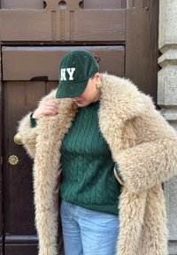 Maglione verde lavorato a trecce sotto un cappotto in pelliccia ecologica marrone chiaro e soffice. Cappello in velluto a coste verde scuro con scritta "NY". Orecchini quadrati dorati.
