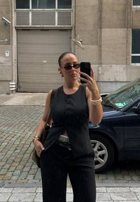 Μαύρο αμάνικο γιλέκο με κουμπιά, συνδυασμένο με φαρδιά μαύρα παντελόνια. Αξεσουάρ περιλαμβάνουν γυαλιά ηλίου, ένα βραχιόλι και μία τσάντα ώμου.