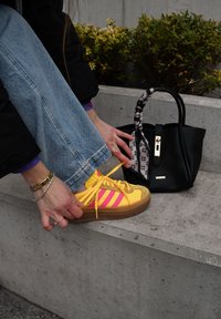 Φωτεινά κίτρινα και ροζ sneakers με καφέ σόλα, συνδυασμένα με ξεθωριασμένο μπλε τζιν. Μια μικρή μαύρη τσάντα με μοτίβο σε κοντινή απόσταση.