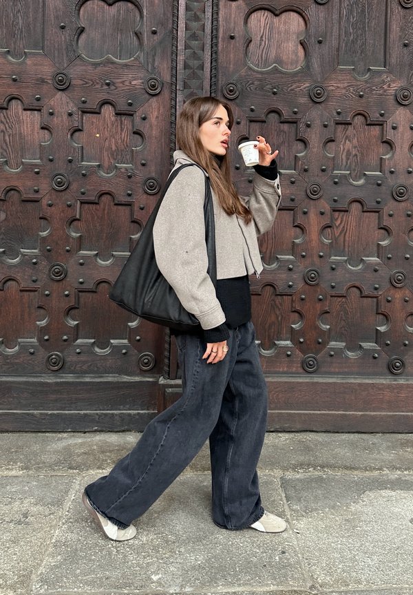 Grey cropped jacket over a black sweater, teamed with wide-leg black jeans and grey sneakers. Carrying a black tote and holding a coffee cup.