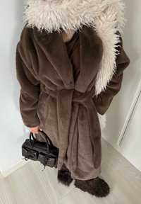 Peignoir en fausse fourrure marron avec un large col châle, taille cintrée et une écharpe beige duveteuse. Tient un petit sac à main noir avec deux poches.