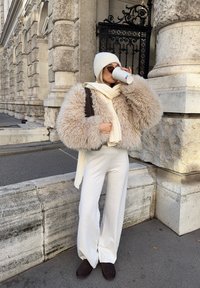 Päällystetty beige tekoturkki, valkoinen neulottu hattu, valkoinen huivi, leveälahkeiset valkoiset housut, ruskeat tossut. Pitelee kuppia, taustalla koristeellinen rakennus.