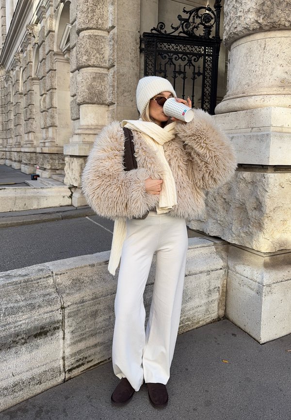 Päällystetty beige tekoturkki, valkoinen neulottu hattu, valkoinen huivi, leveälahkeiset valkoiset housut, ruskeat tossut. Pitelee kuppia, taustalla koristeellinen rakennus.