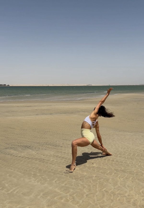 Femme en brassière de sport blanche et short jaune s'étire sur une plage de sable près d'une mer calme sous un ciel dégagé.