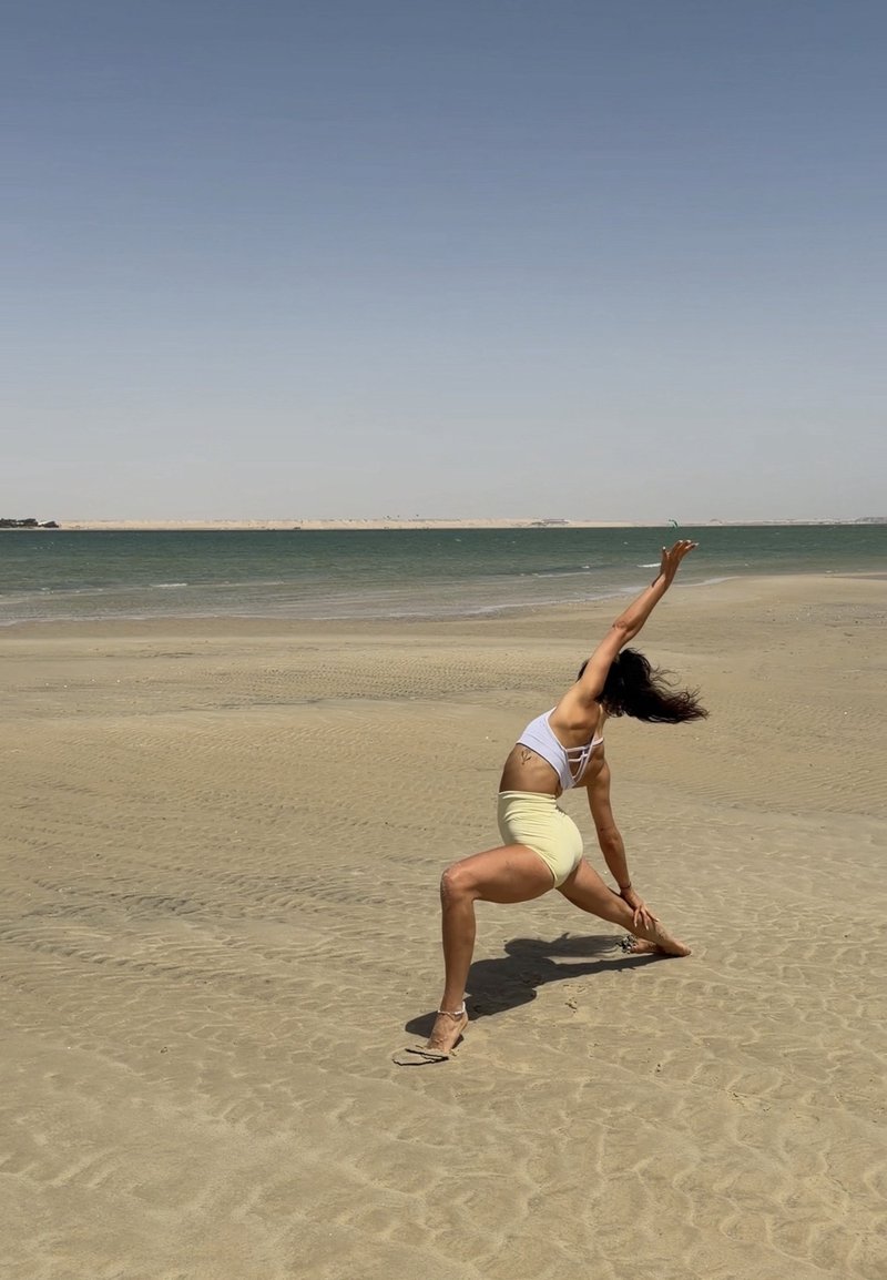 Femme en brassière de sport blanche et short jaune s'étire sur une plage de sable près d'une mer calme sous un ciel dégagé.
