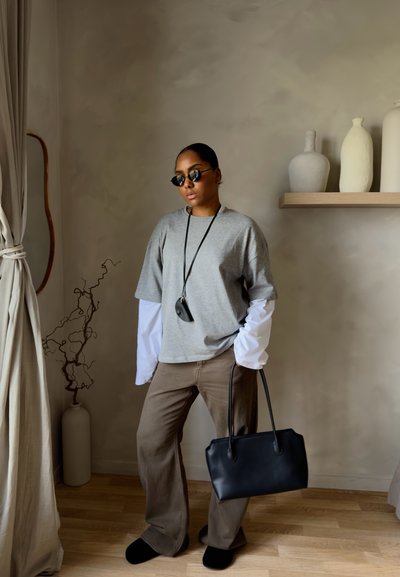 Persona con gafas de sol y prendas superpuestas en tonos gris y blanco, pantalones marrones, sosteniendo un bolso negro, de pie en un interior con una decoración minimalista.