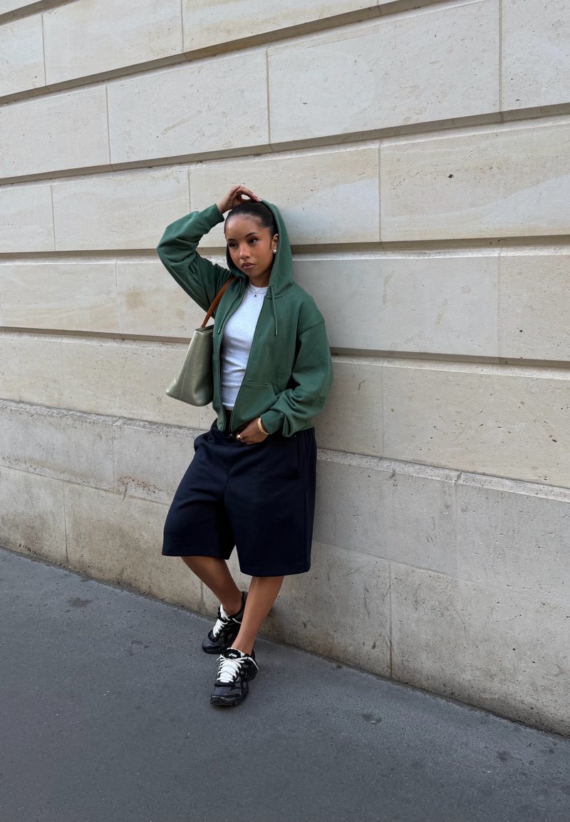 Femme en sweat à capuche vert et short noir appuyée contre un mur en pierre beige, une main sur la tête et portant un sac beige à l'épaule.