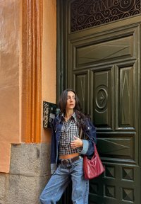 Casaco de ganga com tons de azul sobre uma camisa às riscas, acompanhado de jeans azul claro e uma mala vermelha com detalhes em tachas. Fundo de porta verde.