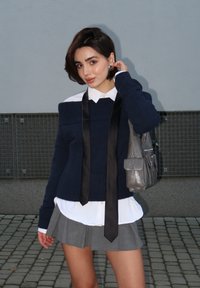 Μπλε πουλόβερ με εκτός ώμου σχέδιο, μαύρη γραβάτα, λευκό πουκάμισο από κάτω, γκρι πλισέ φούστα, μικρή γκρι τσάντα, εξωτερικός χώρος.