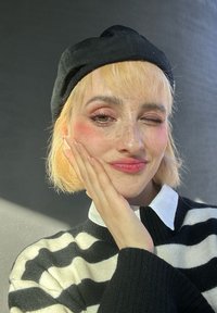 Μοντέλο φοράει μία μαύρη μπερέ και μία ριγέ μαύρο-άσπρη μπλούζα με λευκό γιακά. Έχει κοντά ξανθά μαλλιά και ορατές φακίδες.