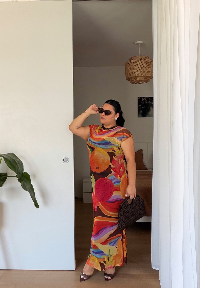 Femme portant une robe colorée à motif de fruits, des lunettes de soleil et un collier noir, tenant un sac noir tissé à l'intérieur près d'une porte coulissante.