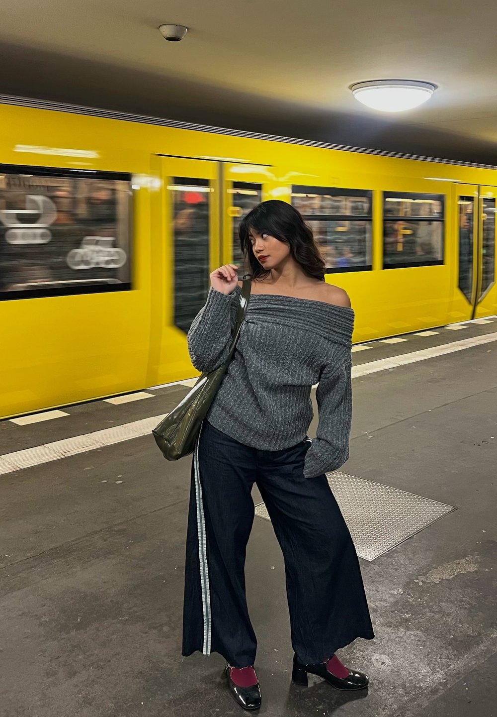 Grijze gebreide trui met blote schouders, brede donkerblauwe jeans met zijstrepen en zwarte hakken. Op de achtergrond staat een gele trein op een station.