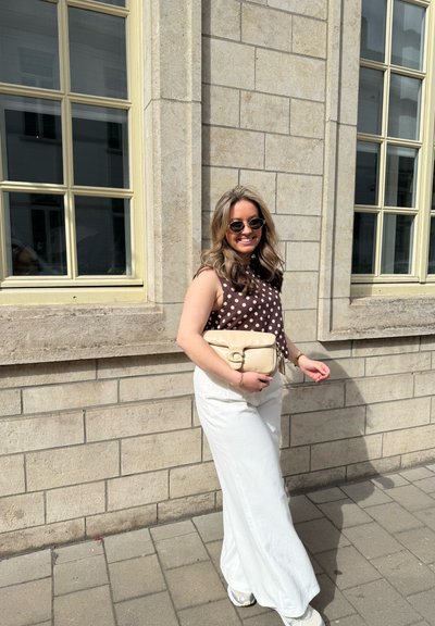 Top de lunares marrones, pantalones amplios blancos, sosteniendo un bolso crema. Gafas de sol redondas, contra una pared de piedra con ventanas. Entorno al aire libre iluminado por el sol.