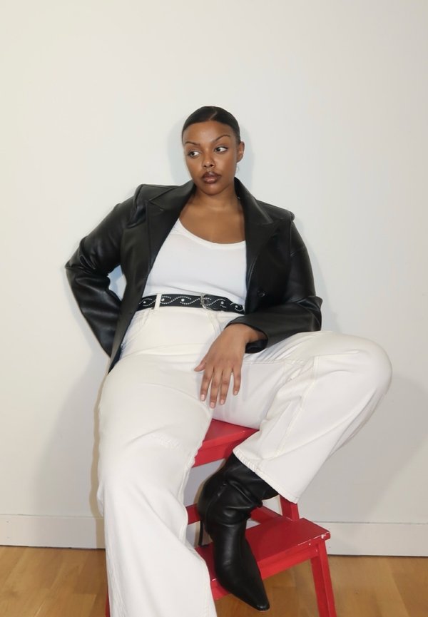 Kvinne sitter på en rød krakk, iført svart skinnjakke, hvit topp, hvite bukser, svarte støvler og et svart belte med sølvdetaljer.