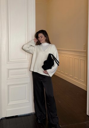 Mujer con suéter blanco y pantalones negros de pie en el interior sosteniendo una bolsa negra, con una mano tocándose el cabello cerca de una puerta panelada.