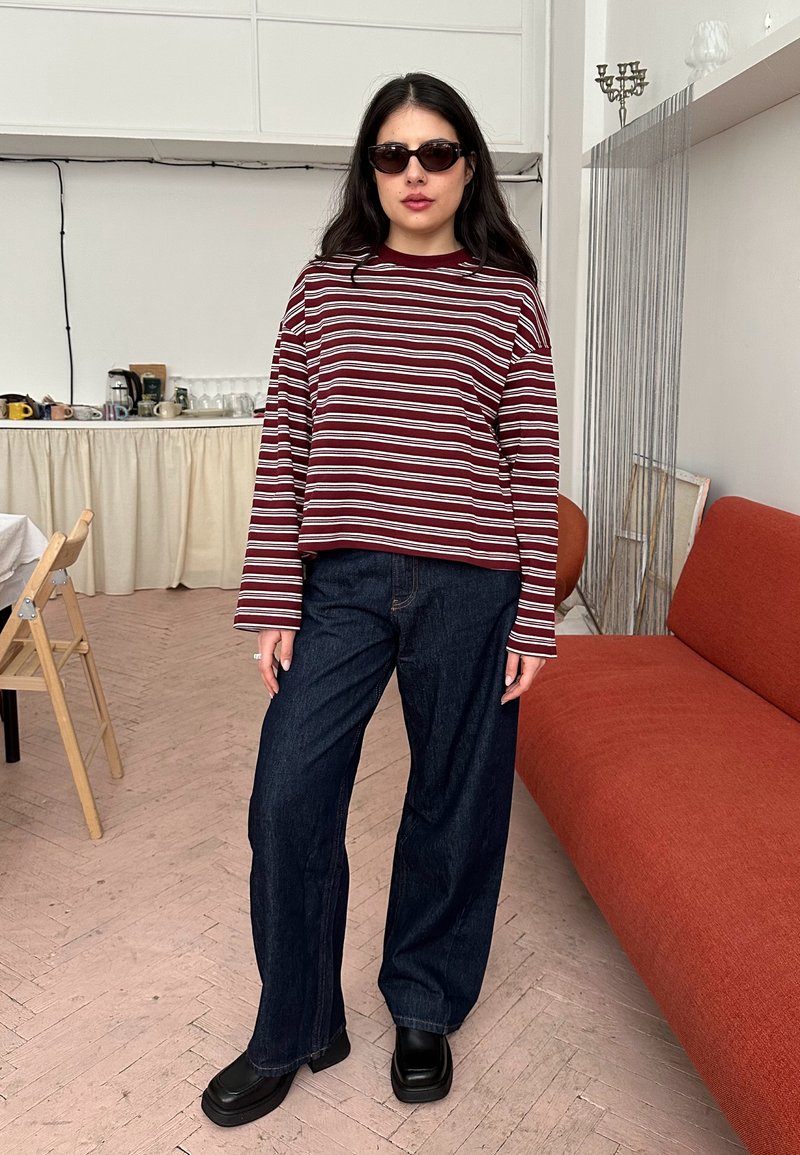 Femme aux longs cheveux bruns porte des lunettes de soleil, un chemisier à rayures bordeaux et blanches à manches longues, un jean large foncé et des chaussures noires à l'intérieur près d'un canapé orange.