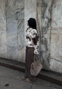 Kvinde i en brun og hvid pelsjakke, brune bukser og støvler, holder en stor beige taske, stående sidelæns på en stengulv ved en marmormur.