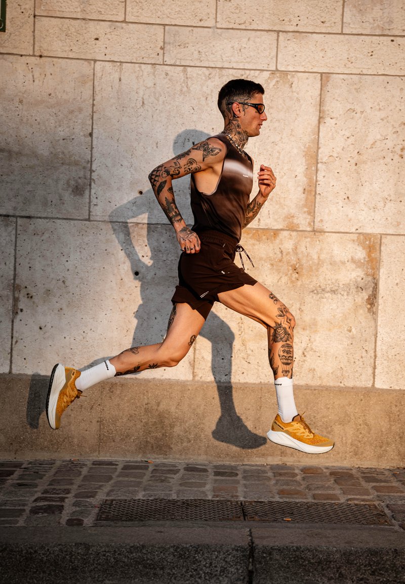 Ténis de corrida amarelos com solas brancas, calções pretos e uma t-shirt de alças em degradê castanho; tatuagens visíveis nos braços e nas pernas; ambiente ao ar livre.